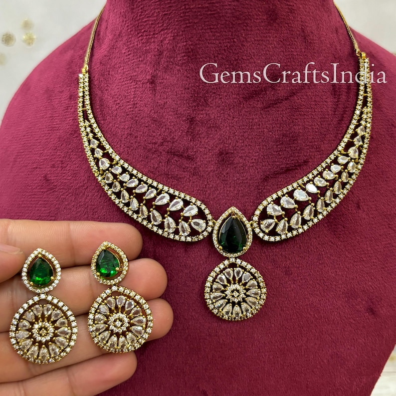 May include: A gold necklace and earrings set featuring a teardrop-shaped green stone and clear stones. The necklace has a curved design and the earrings have a round design.