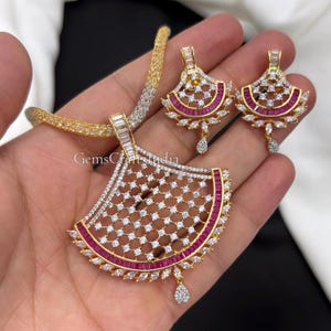 May include: A gold-tone necklace and earring set featuring fan-shaped pendants adorned with clear baguette and round stones, along with vibrant red rectangular stones. The necklace includes a textured chain, and the jewelry displays the text GemsCraftsIndia.