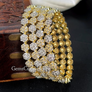May include: Gold-toned bangles with a floral design, encrusted with clear, sparkling stones. The bangles are stacked together, showcasing the intricate details of the jewelry. The image includes the text "GemsCraftsIndia".