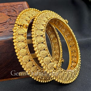 Puede incluir: Dos brazaletes dorados ornamentados con intrincados diseños en relieve. Los brazaletes presentan un patrón de gotas repetido y pequeños detalles de cuentas. Las joyas se exhiben sobre una superficie oscura, mostrando la detallada artesanía.