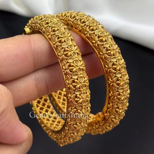 Könnte beinhalten: Zwei goldfarbene Armreifen mit einem detaillierten, erhabenen Blumen- und Blattmuster auf der Außenseite. Das Innere der Armreifen weist ein feines, durchbrochenes Gitterdesign auf. Der Text GemsCraftsIndia ist auf dem Metall sichtbar.