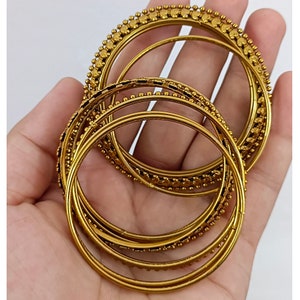 K&ouml;nnte beinhalten: Eine Sammlung goldfarbener Armreifen, einige mit Perlenbesatz, wird gezeigt. Die Armreifen sind kreisf&ouml;rmig und weisen unterschiedliche Designs auf. Das Bild ist vor einem schlichten wei&szlig;en Hintergrund aufgenommen, um die Farbe und Textur des Schmucks hervorzuheben.