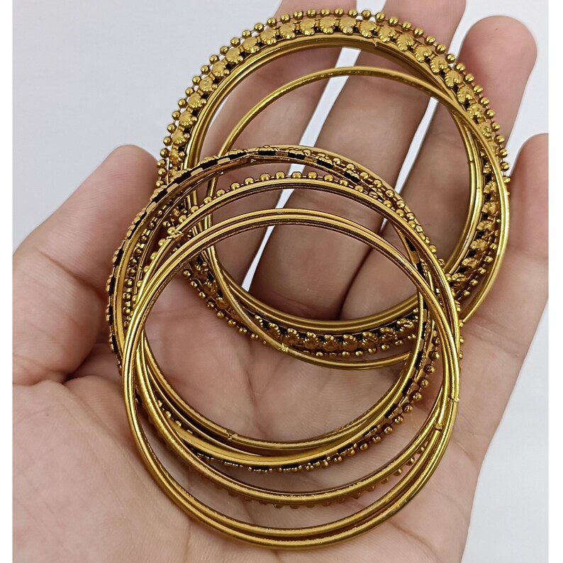 K&ouml;nnte beinhalten: Eine Sammlung goldfarbener Armreifen, einige mit Perlenbesatz, wird gezeigt. Die Armreifen sind kreisf&ouml;rmig und weisen unterschiedliche Designs auf. Das Bild ist vor einem schlichten wei&szlig;en Hintergrund aufgenommen, um die Farbe und Textur des Schmucks hervorzuheben.