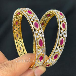 May include: Two gold bangles with a white stone and pink stone design. The bangles are stacked on top of each other.