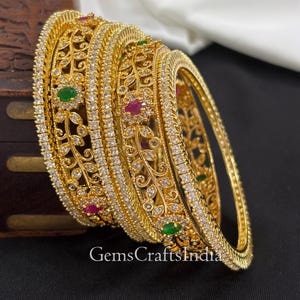 May include: A collection of gold-toned bangles featuring detailed filigree patterns. The bangles are embellished with clear and coloured gemstones, including green and red stones. The text "GemsCraftsIndia" is visible at the bottom of the image.