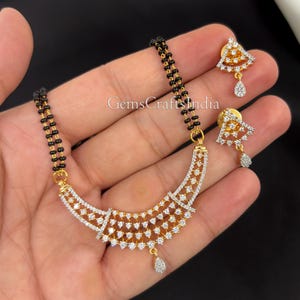May include: A gold-toned necklace and earring set featuring a black beaded chain and a pendant adorned with clear, sparkling stones. The earrings have a triangular design with a dangling teardrop-shaped stone. The text "GemsCraftsIndia" is visible.