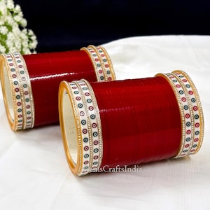 May include: Two red bangles with white and gold decorative bands. The bands feature a repeating pattern of white circles with a black center and a red dot.