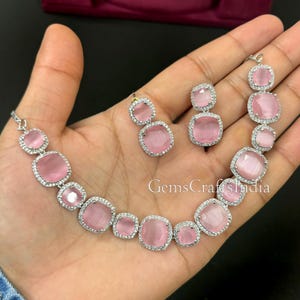 May include: A pink and silver necklace and earrings set with a square-shaped pink stone surrounded by clear rhinestones. The set is on a hand.