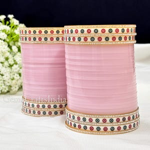 May include: Two pink bangles with gold and white accents. The bangles are decorated with small, colorful circles and rhinestones.