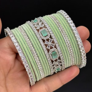 May include: A collection of light green bangles with silver-toned detailing. The bangles have a soft, velvety texture. Some are decorated with clear, sparkling stones and rectangular green stones. The jewellery is shown against a black backdrop.