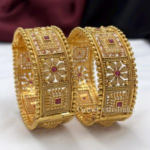 May include: Two gold-toned bangles featuring intricate filigree patterns, floral motifs, and square-shaped accents. Each piece is set with small, deep red stones and clear crystals. The text GemsCraftsIndia is visible on the lower right side.