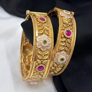 May include: Two gold-tone bangles featuring intricate leaf patterns, embedded pink and green stones, and white enamel flower motifs. The metal surface has a textured finish with raised details. The text GemsCraftsIndia is visible on the lower left.