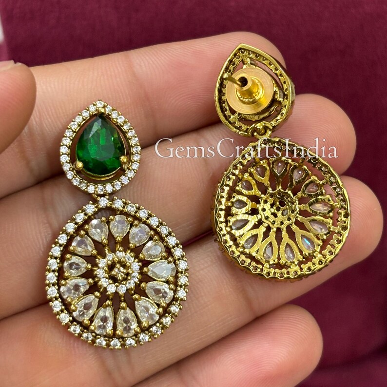 May include: Gold tone dangle earrings featuring a green pear-shaped stone and a round design with clear stones. The earrings are displayed on a hand.