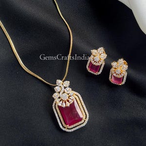 May include: A gold-toned necklace and matching earrings set. The pendant and earrings feature a rectangular, deep red gemstone surrounded by a frame of small, clear stones. The necklace chain is delicate, and the set is displayed on a black surface.