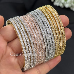 May include: A collection of tennis bracelets in gold, silver, and rose gold tones. Each bracelet is encrusted with small, clear gemstones. The bracelets are stacked together, showcasing their design and color variations. The text "GemsCraftsIndia" is visible.