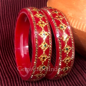 May include: Two red plastic bangles with gold and green decorative accents. The bangles have a geometric pattern with small green stones.