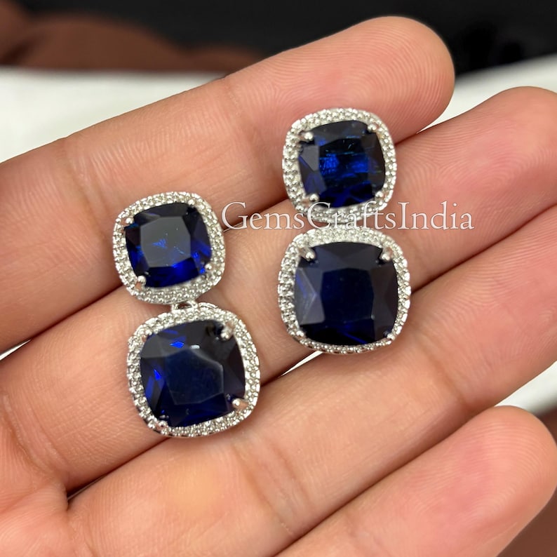 Puede incluir: Un par de pendientes de plata con una gran piedra preciosa cuadrada de color azul oscuro rodeada de piedras preciosas m&aacute;s peque&ntilde;as de color claro. Los pendientes son colgantes y tienen dos piedras preciosas cada uno.