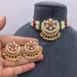 May include: A multi-strand choker necklace and matching stud earrings set featuring white seed pearls, small magenta stones, and green accents. The central crescent-shaped pendant and earrings are adorned with gold-tone metal and dangling pearl beads.
