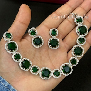 May include: A silver necklace and earrings set with emerald green square-shaped stones surrounded by smaller clear stones. The necklace has a total of 10 stones and the earrings have 2 stones each.