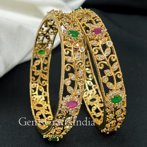 Op de afbeelding: Een paar gouden armbanden met ingewikkelde bloemmotieven, versierd met heldere en groene stenen. De armbanden zijn geplaatst tegen een witte achtergrond.