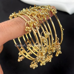 May include: A collection of gold-coloured bangles. Each bangle features small, round, decorative details. The bangles are stacked, highlighting their design and craftsmanship. The text "GemsCraftsIndia" is visible on the bangles.