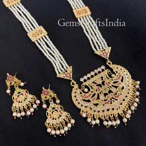May include: A gold-toned necklace and earring set with white pearl strands and intricate floral designs. The necklace features a large pendant with colorful gemstones and dangling pearls. The earrings mirror the pendant's design.