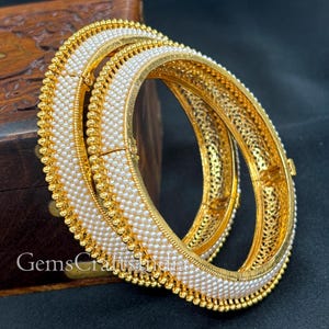 May include: Two gold bangles with white pearl detailing. The inner band features a detailed filigree design. The bangles are set against a dark background. The text "GemsCraftsIndia" is visible.