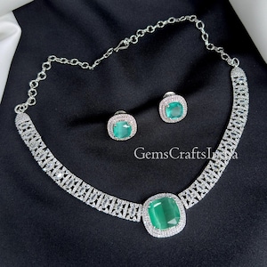 May include: A silver necklace and earring set with emerald-green stones. The necklace has a large, square central stone and a chain. The earrings mirror the design. The set is displayed on a black background. The necklace and earrings are adorned with clear stones.