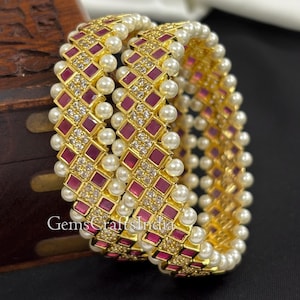 Puede incluir: Dos brazaletes dorados con un patrón repetitivo de cuadrados rojos, pequeñas piedras transparentes y perlas blancas. Los brazaletes tienen una banda dorada y están decorados con el texto "GemsCraftsIndia".