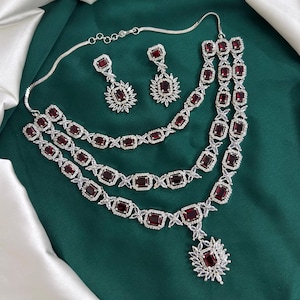 Op de afbeelding: Een zilverkleurige sieradenset met een gelaagde ketting en bijpassende oorbellen. De set is versierd met rechthoekige, dieprode edelstenen en heldere, sprankelende accenten. De ketting heeft een hanger met een grote rode steen. De sieraden worden op een groene achtergrond getoond.