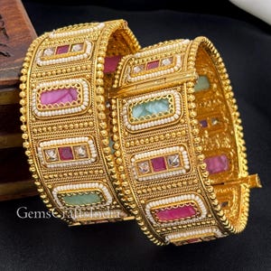 Peut inclure: Deux bracelets rigides en métal doré ornés d'incrustations rectangulaires de pierres roses, rouges et vert clair. Chaque pierre est entourée de petites perles blanches et de détails métalliques dorés. Les bracelets ont un bord perlé.