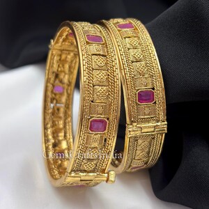May include: Two gold-toned hinged bangles featuring a textured, patterned surface with rectangular, deep pink inlaid stones. The bracelets have a metallic finish with intricate geometric engravings and a visible hinge mechanism on the side.