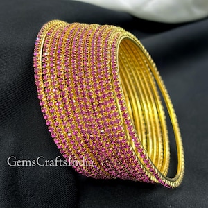 Może przedstawiać: Stos złotych bransoletek inkrustowanych małymi, różowymi kamieniami szlachetnymi. Bransoletki ułożone są w zakrzywionym kształcie, a kamienie szlachetne odbijają światło. Tekst "GemsCraftsIndia" jest widoczny po lewej stronie.