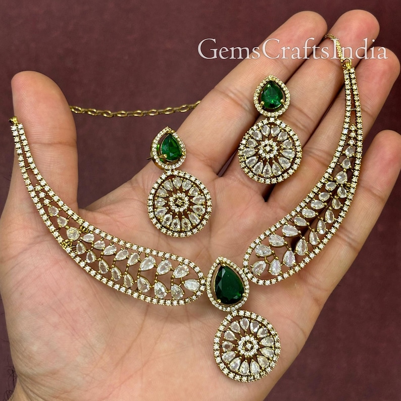 May include: A gold necklace and earrings set with a green stone pendant and clear stones. The necklace is a curved design with a large round pendant.