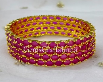 Puede incluir: Un brazalete dorado con tres filas horizontales de piedras ovaladas de color rosa vibrante engastadas en un marco metálico. El texto GemsCraftsIndia es visible en el centro del brazalete, que tiene un patrón repetitivo de piedras.