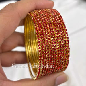 Op de afbeelding: Een set goudkleurige armbanden bezet met kleine, levendige rode edelstenen. De armbanden zijn op elkaar gestapeld, wat hun ingewikkelde ontwerp en de glans van de rode stenen benadrukt. De tekst "GemsCraftsIndia" is zichtbaar.