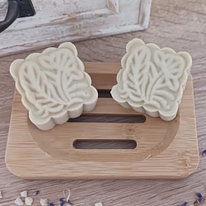 Könnte beinhalten: Zwei weiße Seifenstücke mit einem Blattdesign auf einem Holzseifenhalter.