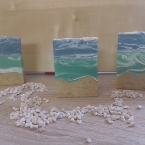 Puede incluir: Tres barras de jab&oacute;n hechas a mano con un dise&ntilde;o de olas de mar azul y verde. Las barras de jab&oacute;n est&aacute;n sobre una superficie de madera con guijarros blancos esparcidos a su alrededor. Las barras de jab&oacute;n tienen el texto "Soap & Sea" en una de las barras.