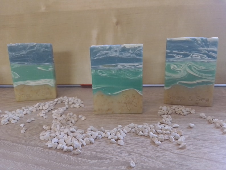 Puede incluir: Tres barras de jab&oacute;n hechas a mano con un dise&ntilde;o de olas de mar azul y verde. Las barras de jab&oacute;n est&aacute;n sobre una superficie de madera con guijarros blancos esparcidos a su alrededor. Las barras de jab&oacute;n tienen el texto "Soap & Sea" en una de las barras.