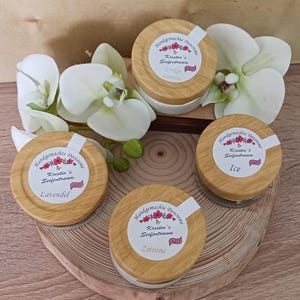 Könnte beinhalten: Vier kleine Glasgefäße mit Holzdeckeln. Jedes Gefäß hat ein weißes Etikett mit einem floralen Design und Text, der "Handgemachte Deocreme" und "Kristin's Seifentraum" lautet. Die Gefäße sind mit den Düften "Zitrone", "Lavendel", "Eis" und "Gokyo Comette" beschriftet.