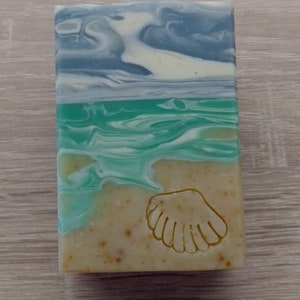 Könnte beinhalten: Eine Seife mit Muschel-Design. Die Seife ist in Schichten aus Blau, Grün und Weiß geschichtet, die an eine Strandszene erinnern. Die unterste Schicht ist hellbraun mit einem Muschelabdruck.