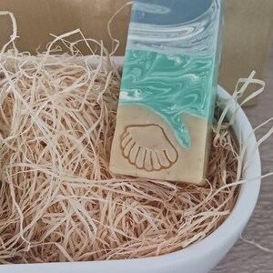 Puede incluir: Una pastilla de jab&oacute;n artesanal con un dise&ntilde;o de concha. El jab&oacute;n est&aacute; en capas de tonos azules, verdes y beige. Est&aacute; sentado en un taz&oacute;n de cer&aacute;mica blanco lleno de papel triturado.