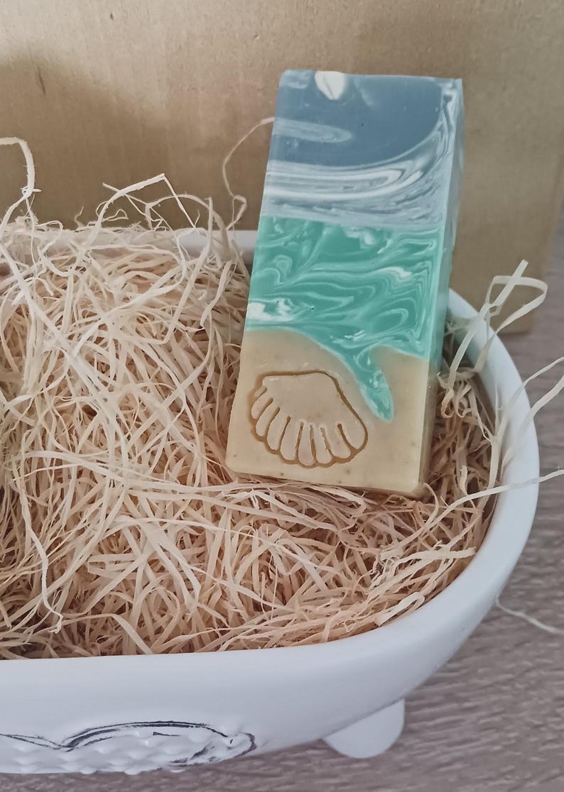 Puede incluir: Una pastilla de jab&oacute;n artesanal con un dise&ntilde;o de concha. El jab&oacute;n est&aacute; en capas de tonos azules, verdes y beige. Est&aacute; sentado en un taz&oacute;n de cer&aacute;mica blanco lleno de papel triturado.
