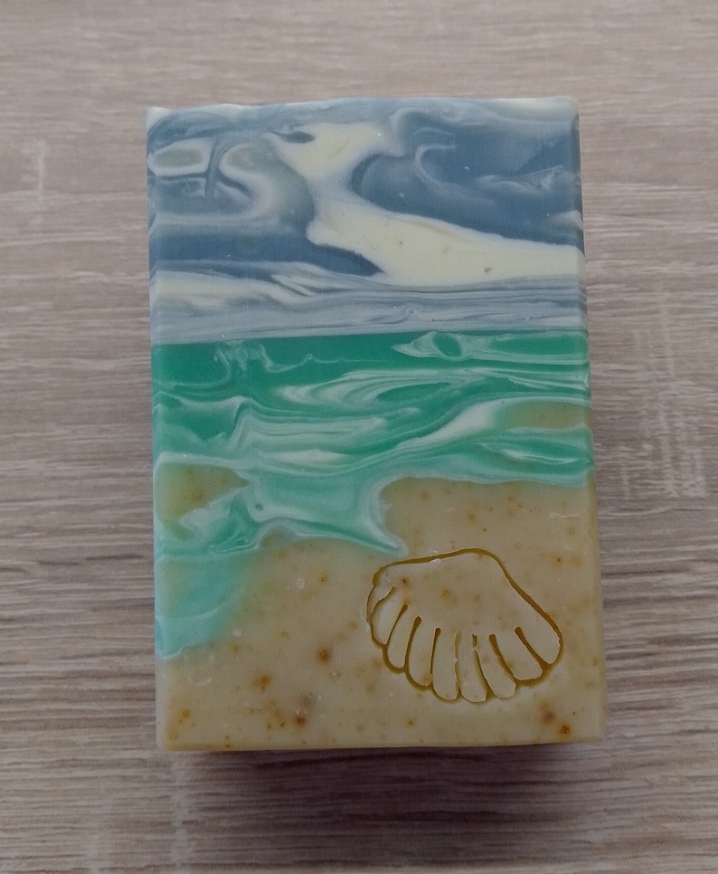 Puede incluir: Una pastilla de jab&oacute;n con un dise&ntilde;o de concha marina. El jab&oacute;n est&aacute; en capas con remolinos azules, verdes y blancos, que se asemejan a una escena de playa.