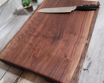 May include: A rectangular, dark brown wooden cutting board with a live edge. A stainless steel knife with a black handle rests on the board. Two small potted succulents are in the background. The board is approximately 18 inches long.