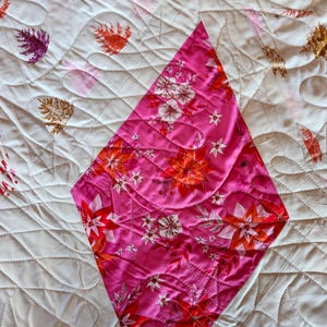 May include: A quilted textile featuring a diamond-shaped section with a floral pattern in shades of pink, red, and white. The surrounding fabric is white with a repeating leaf pattern in various colors. The quilt has a stitched, textured design.