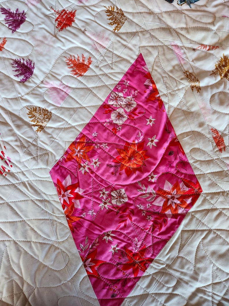 May include: A quilted textile featuring a diamond-shaped section with a floral pattern in shades of pink, red, and white. The surrounding fabric is white with a repeating leaf pattern in various colors. The quilt has a stitched, textured design.