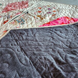 May include: A quilted textile with a cream-colored top featuring a patchwork design with pink and red floral patterns and whimsical illustrations. The underside is a textured gray fabric with a quilted pattern.