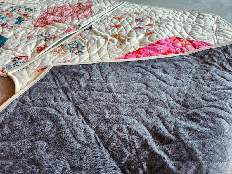 May include: A quilted textile with a cream-colored top featuring a patchwork design with pink and red floral patterns and whimsical illustrations. The underside is a textured gray fabric with a quilted pattern.