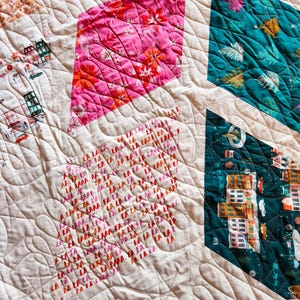 May include: A quilted textile featuring a patchwork design with various geometric shapes. The quilt showcases vibrant colors, including pink floral patterns, teal, and a cream-colored background. The text "LA LA LA" is visible on one of the patches.